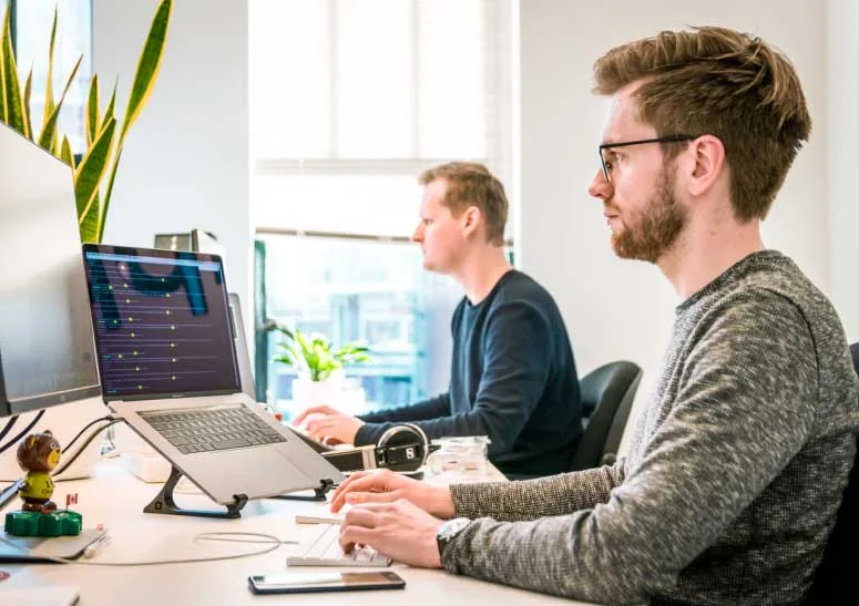 "Digital marketers seated side by side at a spacious table, diligently working on their laptops.
